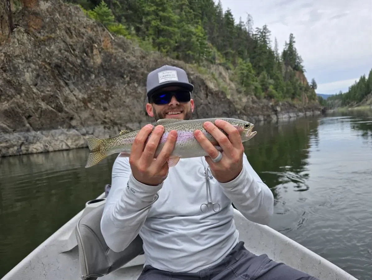 Austin Lewis, founder of GuidedFloats, holding a rainbow trout from a drift boat beneath a canyon wall