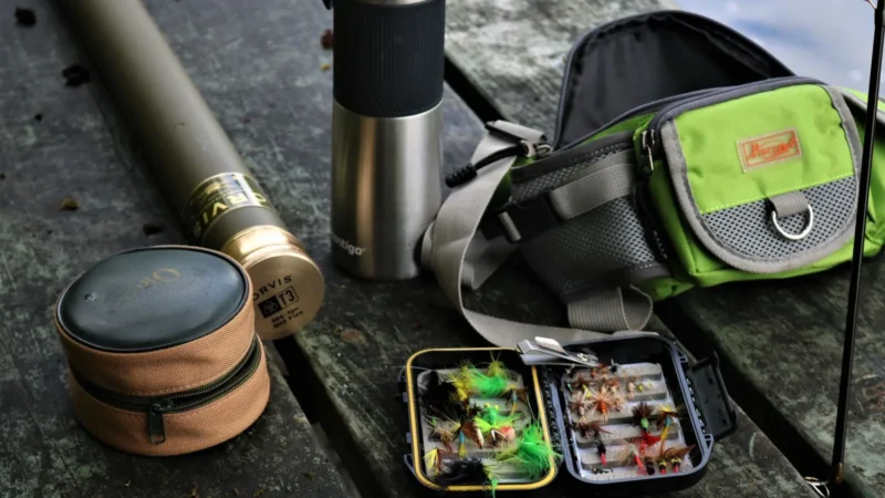 A fly rod, reel case, fly boxes, thermos and sling pack laid out on a rock