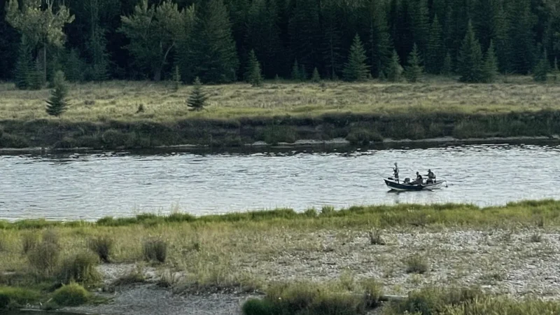 A drift boat with three anglers on a wide river below a forested bank
