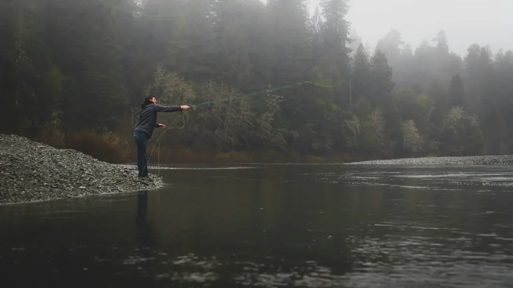 A fly angler casting from a gravel bar into a fog-covered river on a cold morning