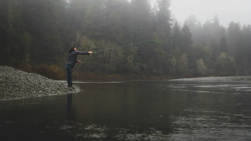 A fly angler casting from a gravel bar into a fog-covered river on a cold morning