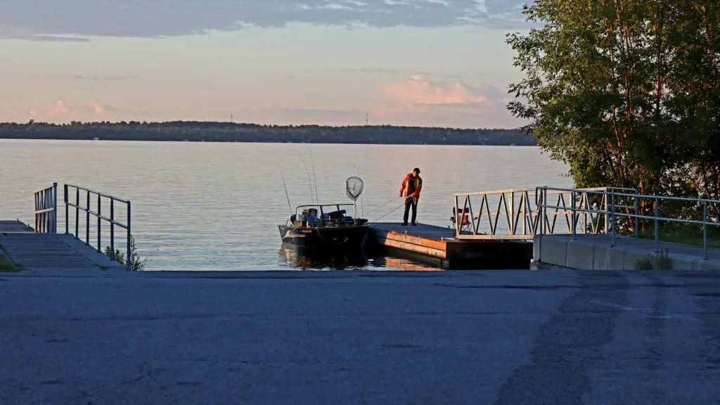 An angler on a dock beside a moored boat on a calm reservoir
