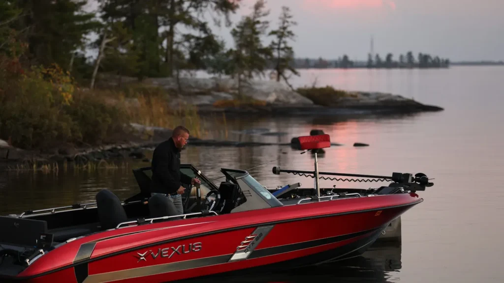 An angler standing in a bass boat along a wooded reservoir shoreline